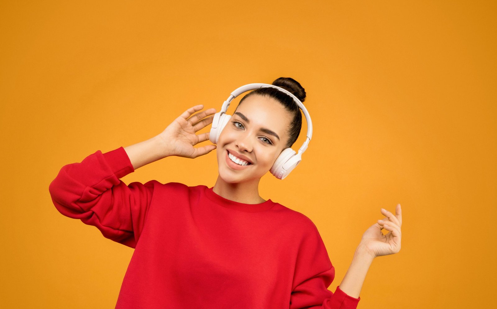 Smiling woman in red sweater enjoying music with wireless headphones against orange background.